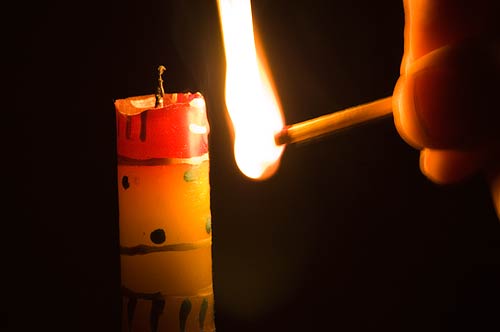 photo: a lighted match, blazing intently, approaches a colorful unlit candle.