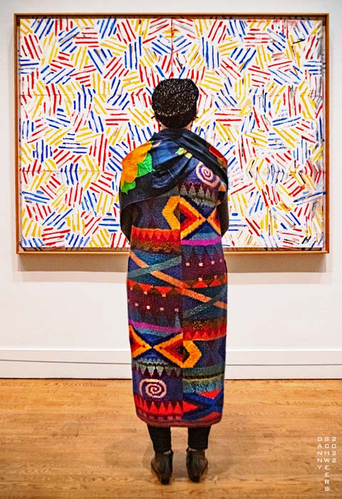 Photo of a colorfully-attired woman standing in front of the painting Corpse and Mirror II by Jasper Johns, photo copyright 2022 by Danny N. Schweers